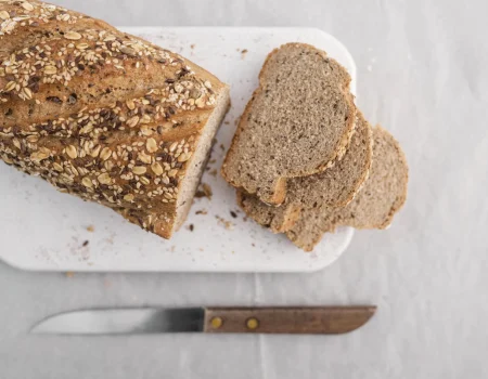 pão caseiro sem glutem com grãos em uma tábua branca sobre uma mesa branca com uma faca ao lado